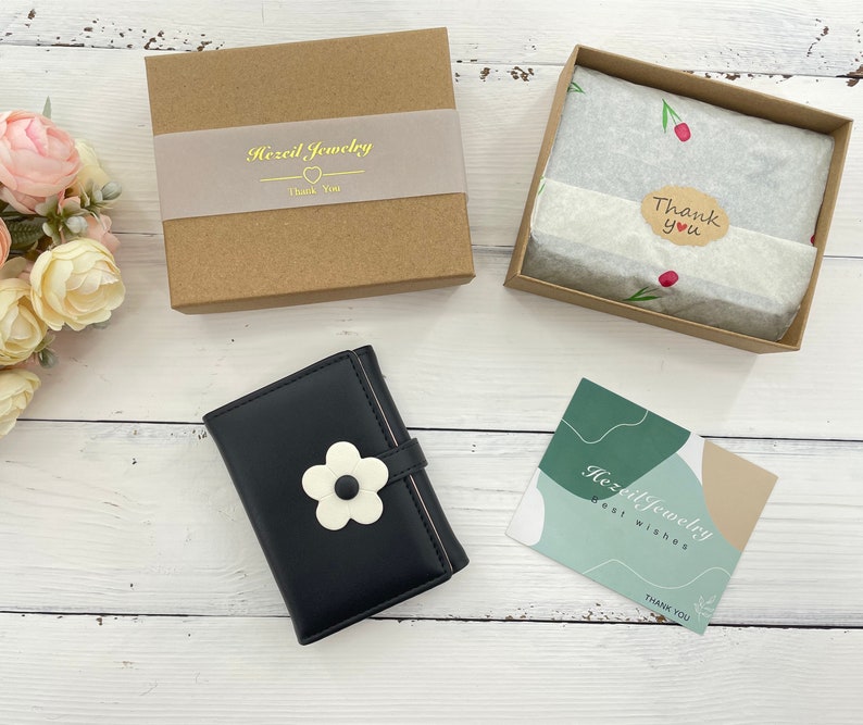 Pu&ograve; includere: Un portafoglio in pelle nera con un motivo a fiore bianco e il testo "Hezi Jewelry" su un biglietto bianco. Il portafoglio &egrave; su una superficie di legno bianco. Il biglietto dice anche "Auguri" e "Grazie".