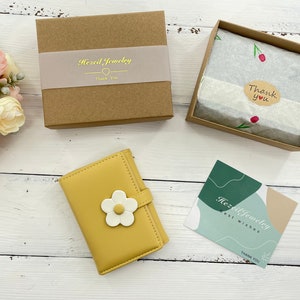 Pu&ograve; includere: Un portafoglio giallo con un design a fiore bianco. Il portafoglio &egrave; chiuso e si trova in una scatola marrone con il testo "Hazel Jewelry" e un biglietto di ringraziamento. C'&egrave; anche un piccolo biglietto con il testo "Hazel Jewelry Best Wishes Thank You".