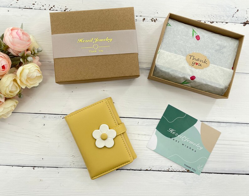 Pu&ograve; includere: Un portafoglio giallo con un design a fiore bianco. Il portafoglio &egrave; chiuso e si trova in una scatola marrone con il testo "Hazel Jewelry" e un biglietto di ringraziamento. C'&egrave; anche un piccolo biglietto con il testo "Hazel Jewelry Best Wishes Thank You".
