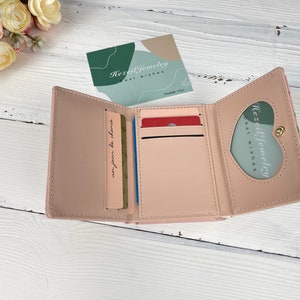 Pu&ograve; includere: Un portafoglio rosa a tre pieghe con una finestra a forma di cuore sul davanti. Il portafoglio &egrave; aperto e mostra tre slot per carte e un vano per contanti. Il testo "Hezil Jewelry Best Wishes" &egrave; visibile all'interno della finestra.