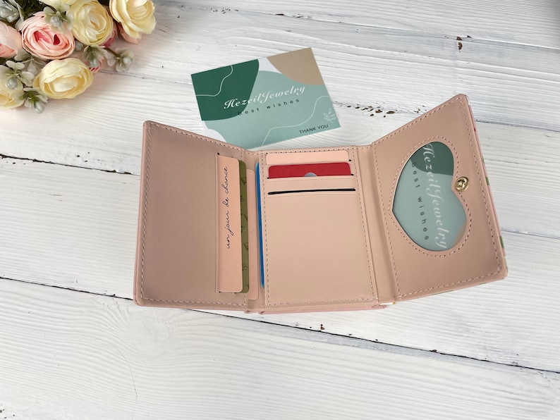 Pu&ograve; includere: Un portafoglio rosa a tre pieghe con una finestra a forma di cuore sul davanti. Il portafoglio &egrave; aperto e mostra tre slot per carte e un vano per contanti. Il testo "Hezil Jewelry Best Wishes" &egrave; visibile all'interno della finestra.