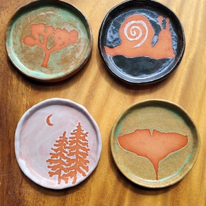 May include: Four small, round ceramic dishes with hand-painted designs. One features a tree, another a snail, a third has trees and a crescent moon, and the last a leaf. Each dish has a different colour scheme.