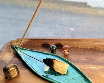 May include: A glossy teal ceramic incense holder shaped like a leaf, featuring a thin incense stick, a piece of charred palo santo wood, a tiger's eye stone, and two small mushroom-shaped stone carvings resting on a smooth wooden surface.