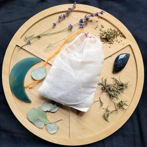 May include: A wooden tray with divided sections holds various items. A white cloth bag with an orange drawstring sits in the center. Other items include sprigs of lavender, dried herbs, a green crescent-shaped stone, and a dark blue stone.