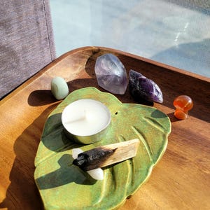 May include: A green leaf-shaped ceramic dish with a white candle, a small piece of burnt palo santo wood, and several amethyst crystals on a wooden tray.