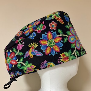 May include: A black surgical scrub cap with a vibrant floral and bird pattern. The design features colourful flowers and birds in shades of red, blue, yellow, and green. The cap has a drawstring for an adjustable fit.
