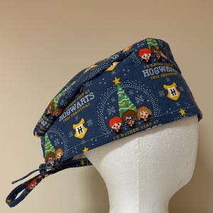 May include: A blue scrub cap featuring a repeating pattern of cartoon characters, Christmas trees, and the text "I'd rather stay at Hogwarts this Christmas." The cap has a tie closure and a yellow Hogwarts crest.