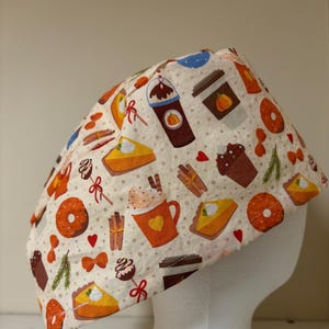 May include: A beige surgical scrub cap featuring a repeating pattern of fall-themed illustrations. The design includes images of pumpkin spice lattes, donuts, pumpkin pie slices, cupcakes, and cinnamon sticks. The cap is designed to fit the head.