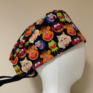 May include: A black surgical scrub cap with a vibrant pattern of Muppet characters. The cap is tied at the back and is shown on a white mannequin head. The Muppet faces include Kermit, Miss Piggy, Fozzie Bear, and other characters.