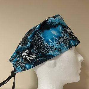 May include: A blue and black patterned scrub cap with a castle design and the words "Harry Potter" in white. The cap has black ties for adjustment and is displayed on a white mannequin head.
