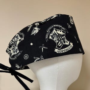 May include: Black scrub cap featuring a repeating pattern of white Hogwarts crests, stars, and other symbols. The cap has a tie closure and is designed for medical use. The cap is made of a soft, durable fabric.