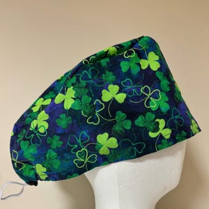 Peut inclure: Une toque de chirurgien à motifs de trèfles verts et bleus. Le bonnet est en tissu et possède un élastique pour un ajustement sûr. Les trèfles sont de différentes nuances de vert, sur un fond bleu foncé.