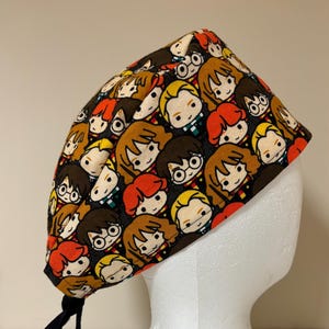 May include: A black surgical scrub cap featuring a repeating pattern of cartoon characters with glasses and various hair colors. The cap has a black tie and is displayed on a white mannequin head.