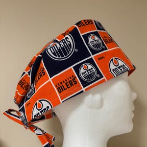 May include: A blue and orange surgical scrub hat with a repeating pattern of the Edmonton Oilers NHL hockey team logo. The hat has ties on the side.
