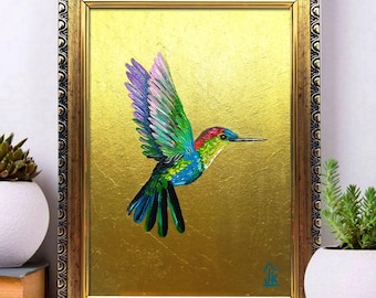 Pode incluir: Uma pintura emoldurada de um beija-flor em voo contra um fundo dourado sólido. O pássaro apresenta uma plumagem vibrante em tons de verde, azul e vermelho. A obra de arte está inserida numa moldura metálica escura e ornamentada com padrão.