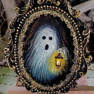 May include: A framed miniature painting of a ghost holding a lantern. The artwork features a white ghost with a surprised expression, set against a dark background. The ornate frame is black and gold, displayed on a small wooden easel.
