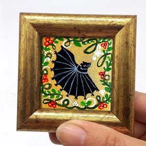 Peut inclure: Petite peinture encadrée représentant une chauve-souris noire aux ailes déployées sur fond doré. La chauve-souris est entourée de vignes vertes et de fleurs rouges et blanches. Le tableau est dans un cadre doré.
