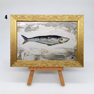 Puede incluir: Una peque&ntilde;a pintura de un pez plateado sobre un fondo blanco con un cielo texturizado gris y blanco. La pintura est&aacute; enmarcada en un marco dorado y se muestra en un peque&ntilde;o caballete de madera. La pintura est&aacute; firmada "JK" en la esquina inferior derecha.
