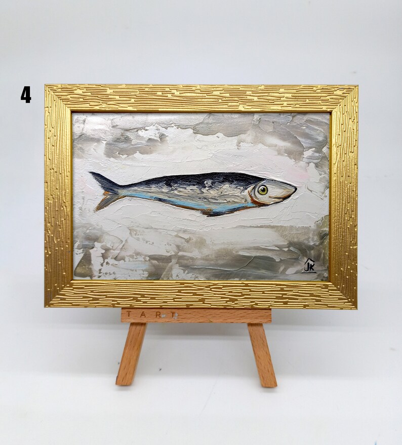 Puede incluir: Una peque&ntilde;a pintura de un pez plateado sobre un fondo blanco con un cielo texturizado gris y blanco. La pintura est&aacute; enmarcada en un marco dorado y se muestra en un peque&ntilde;o caballete de madera. La pintura est&aacute; firmada "JK" en la esquina inferior derecha.