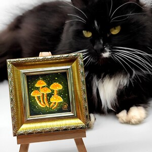 May include: A small framed painting of a cluster of orange mushrooms on a green background with a black cat sitting in the foreground. The painting is on a wooden easel.