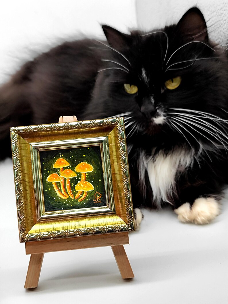May include: A small framed painting of a cluster of orange mushrooms on a green background with a black cat sitting in the foreground. The painting is on a wooden easel.
