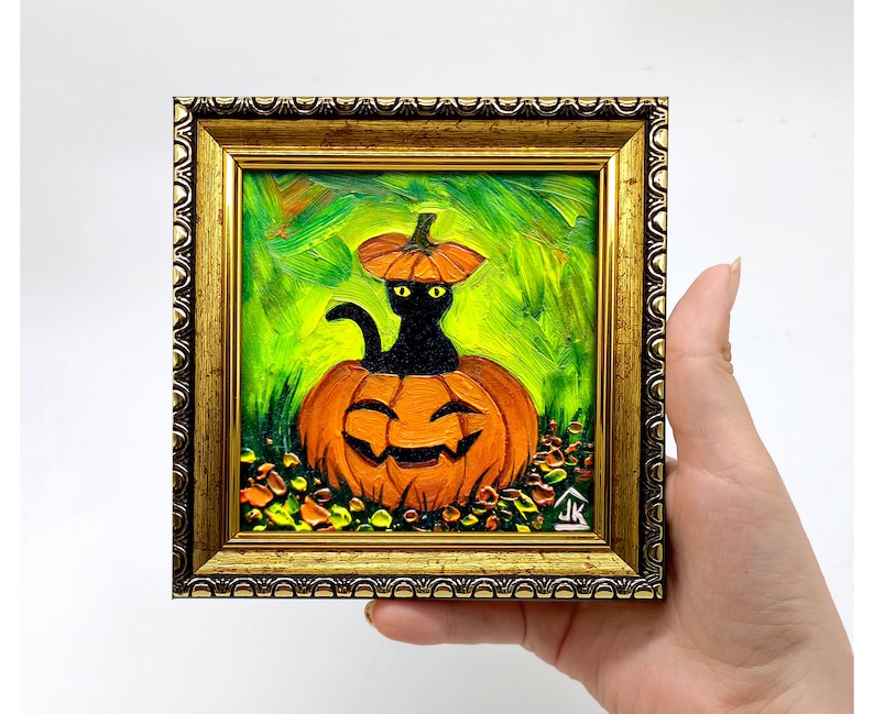 May include: A framed Halloween-themed painting. The artwork depicts a black cat inside an orange jack-o'-lantern, with a smaller pumpkin hat. The background is green, and the frame is gold. The painting is held by a hand.