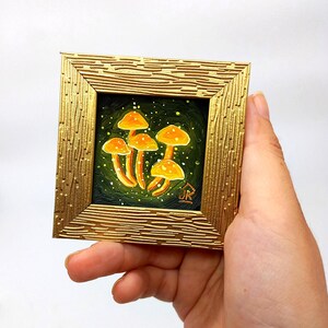 May include: A small framed painting of a cluster of orange mushrooms with white spots on a dark green background. The mushrooms are painted in a whimsical style with a glowing effect. The frame is gold with a textured surface.