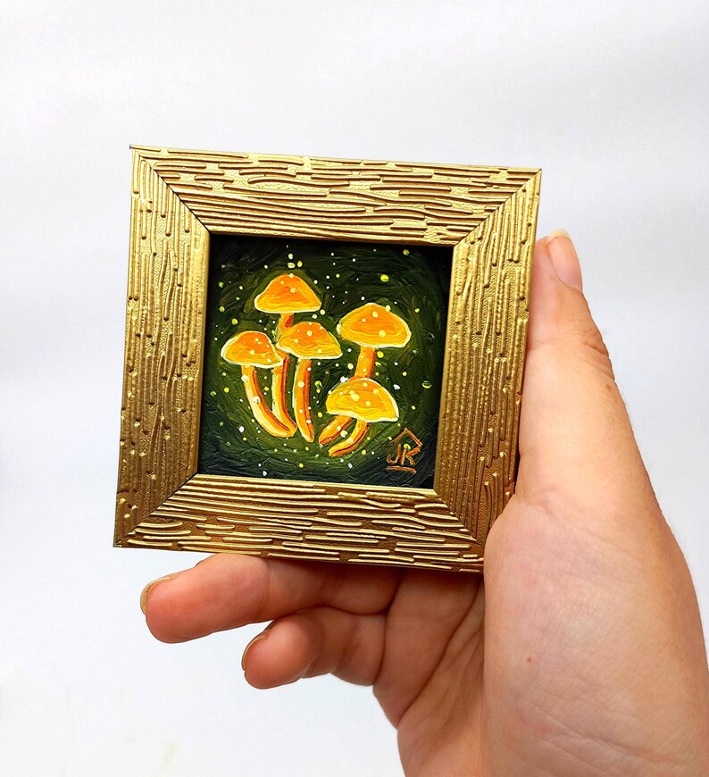 May include: A small framed painting of a cluster of orange mushrooms with white spots on a dark green background. The mushrooms are painted in a whimsical style with a glowing effect. The frame is gold with a textured surface.