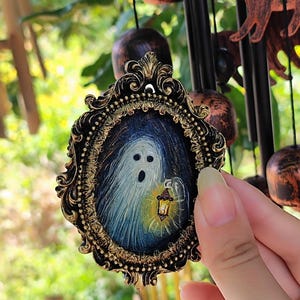 May include: An oval pendant with a detailed gold frame, featuring a Halloween ghost design. The ghost is holding a lantern, set against a dark blue background. The pendant is a decorative item, possibly for a necklace.
