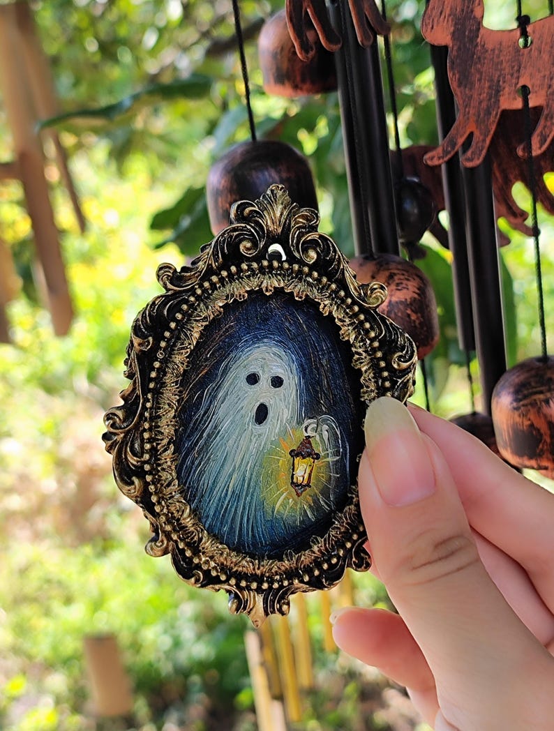 May include: An oval pendant with a detailed gold frame, featuring a Halloween ghost design. The ghost is holding a lantern, set against a dark blue background. The pendant is a decorative item, possibly for a necklace.