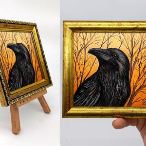 Pode incluir: Uma obra de arte emoldurada com uma pintura detalhada de um corvo preto contra um fundo laranja e castanho com galhos de &aacute;rvores nus. A obra de arte est&aacute; em uma moldura dourada e &eacute; exibida em um pequeno cavalete de madeira.