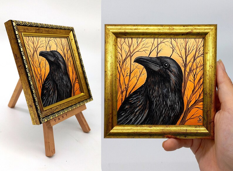 Pode incluir: Uma obra de arte emoldurada com uma pintura detalhada de um corvo preto contra um fundo laranja e castanho com galhos de &aacute;rvores nus. A obra de arte est&aacute; em uma moldura dourada e &eacute; exibida em um pequeno cavalete de madeira.