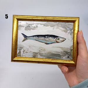 Puede incluir: Una pintura enmarcada de un pez plateado con una franja azul sobre un fondo blanco con pinceladas grises y marrones. El marco es dorado y tiene una peque&ntilde;a firma en la esquina inferior derecha.