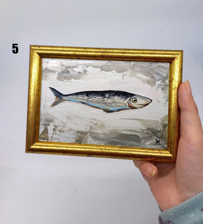 Puede incluir: Una pintura enmarcada de un pez plateado con una franja azul sobre un fondo blanco con pinceladas grises y marrones. El marco es dorado y tiene una peque&ntilde;a firma en la esquina inferior derecha.