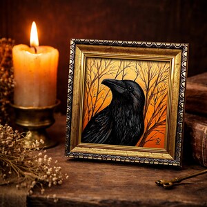 Pode incluir: Uma obra de arte emoldurada com um corvo preto contra um fundo laranja e amarelo com galhos de &aacute;rvores nus. A obra de arte est&aacute; em uma moldura dourada ornamentada. Uma vela acesa e flores secas est&atilde;o no fundo, criando um tema g&oacute;tico ou m&iacute;stico.
