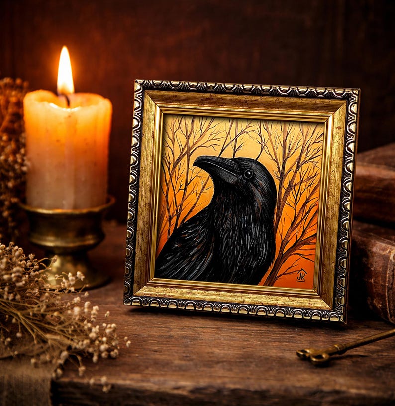 Pode incluir: Uma obra de arte emoldurada com um corvo preto contra um fundo laranja e amarelo com galhos de &aacute;rvores nus. A obra de arte est&aacute; em uma moldura dourada ornamentada. Uma vela acesa e flores secas est&atilde;o no fundo, criando um tema g&oacute;tico ou m&iacute;stico.