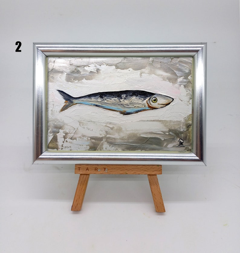 Puede incluir: Una peque&ntilde;a pintura enmarcada de un pez plateado sobre un fondo blanco con un efecto de textura de pinceladas. La pintura est&aacute; en un caballete con la palabra "TART" escrita en &eacute;l.