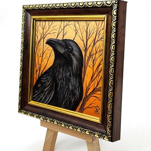 Pode incluir: Pintura emoldurada de um corvo preto com um fundo laranja e castanho de &aacute;rvores despidas. A obra de arte &eacute; exibida num pequeno cavalete de madeira. A moldura castanha escura com detalhes dourados real&ccedil;a as cores e texturas ricas.
