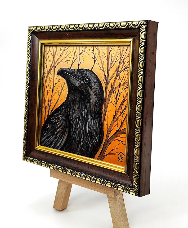 Pode incluir: Pintura emoldurada de um corvo preto com um fundo laranja e castanho de &aacute;rvores despidas. A obra de arte &eacute; exibida num pequeno cavalete de madeira. A moldura castanha escura com detalhes dourados real&ccedil;a as cores e texturas ricas.