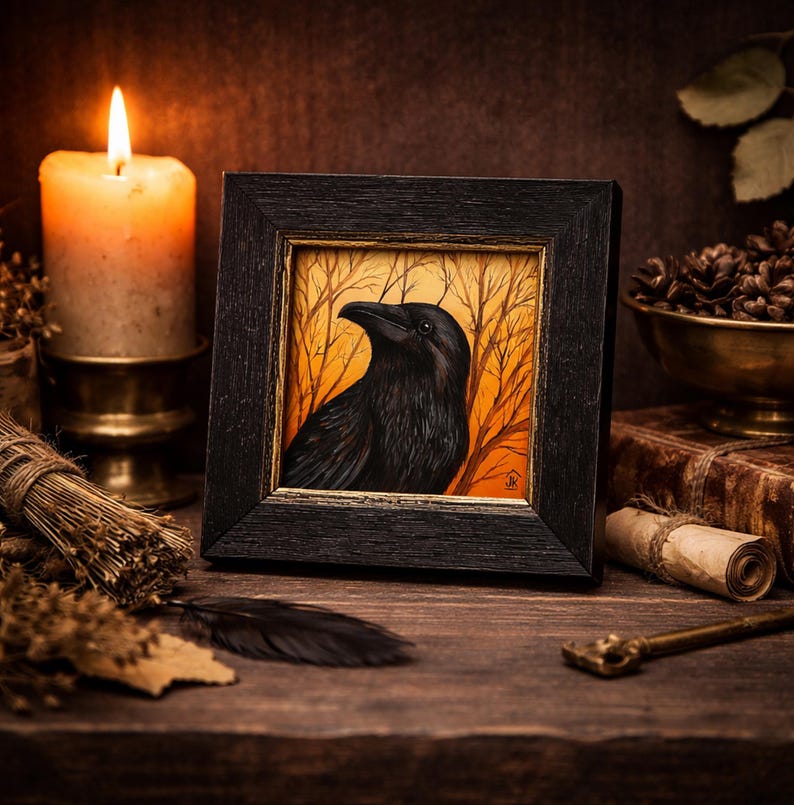 Pode incluir: Uma obra de arte emoldurada com um corvo preto contra um fundo laranja e castanho com &aacute;rvores nuas. O corvo &eacute; o ponto focal, emoldurado em madeira escura. Uma vela acesa, ervas secas e uma pena est&atilde;o em uma superf&iacute;cie de madeira.
