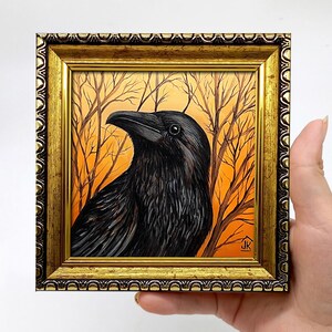 Pode incluir: Pintura emoldurada de um corvo preto com penas detalhadas, contra um fundo de &aacute;rvores nuas e c&eacute;u laranja. A obra de arte est&aacute; fechada em uma moldura dourada ornamentada, criando uma est&eacute;tica vintage.