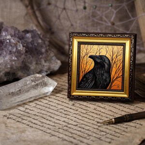Pode incluir: Uma obra de arte emoldurada com um corvo preto contra um fundo laranja e amarelo com &aacute;rvores nuas. A moldura &eacute; dourada e castanha. A obra de arte &eacute; exibida em papel vintage com um cristal de quartzo transparente e cristais de ametista.