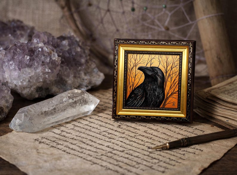 Pode incluir: Uma obra de arte emoldurada com um corvo preto contra um fundo laranja e amarelo com &aacute;rvores nuas. A moldura &eacute; dourada e castanha. A obra de arte &eacute; exibida em papel vintage com um cristal de quartzo transparente e cristais de ametista.