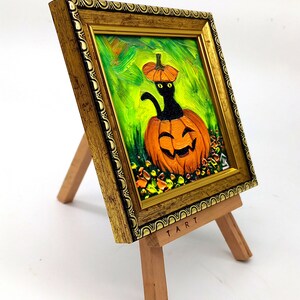 Op de afbeelding: Een olieverfschilderij in een versierde gouden lijst op een kleine houten ezel. Het kunstwerk toont een zwarte kat met gele ogen die uit een uitgesneden oranje pompoen komt tegen een levendige groene achtergrond. Het woord TART staat op de ezel.