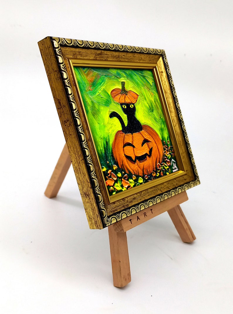 Op de afbeelding: Een olieverfschilderij in een versierde gouden lijst op een kleine houten ezel. Het kunstwerk toont een zwarte kat met gele ogen die uit een uitgesneden oranje pompoen komt tegen een levendige groene achtergrond. Het woord TART staat op de ezel.