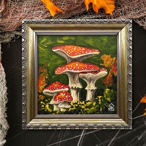 May include: A framed painting of three red and white mushrooms with white spots on a green background. The mushrooms are in a forest setting with leaves and other foliage. The painting is in a silver frame with a decorative border.