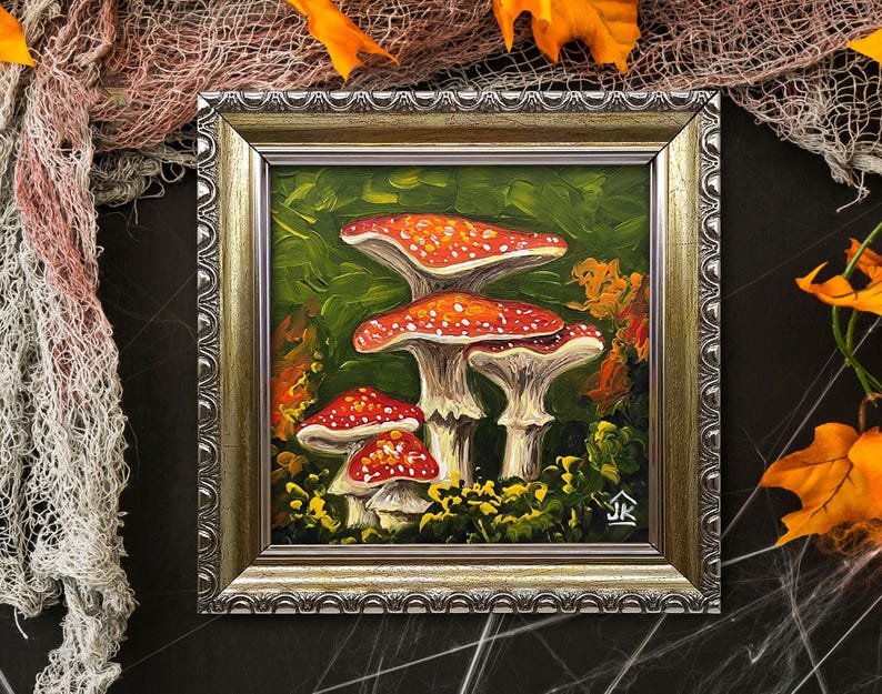 May include: A framed painting of three red and white mushrooms with white spots on a green background. The mushrooms are in a forest setting with leaves and other foliage. The painting is in a silver frame with a decorative border.