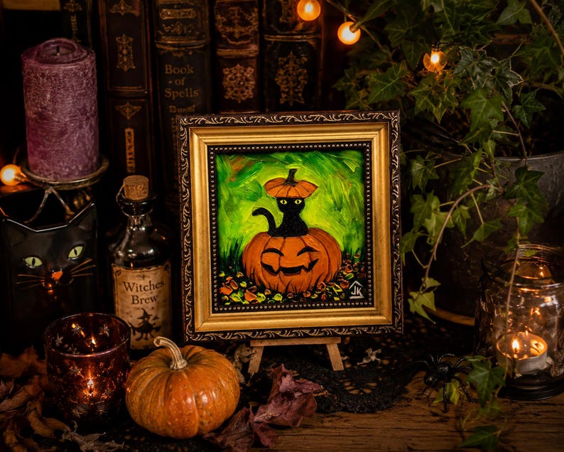 Op de afbeelding: Een ingelijst olieverfschilderij met een zwarte kat die in een uitgesneden oranje pompoen zit en een kleine pompoen als hoed draagt. Het kunstwerk is geplaatst tegen een levendige groene achtergrond met herfstbladeren aan de basis.