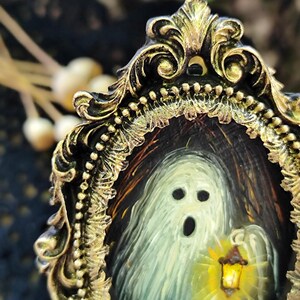 May include: A small painting of a ghost with a lantern, framed in an elaborate gold-coloured frame. The ghost is rendered in white and grey, with a black open mouth and eye sockets. The frame features detailed, decorative elements.