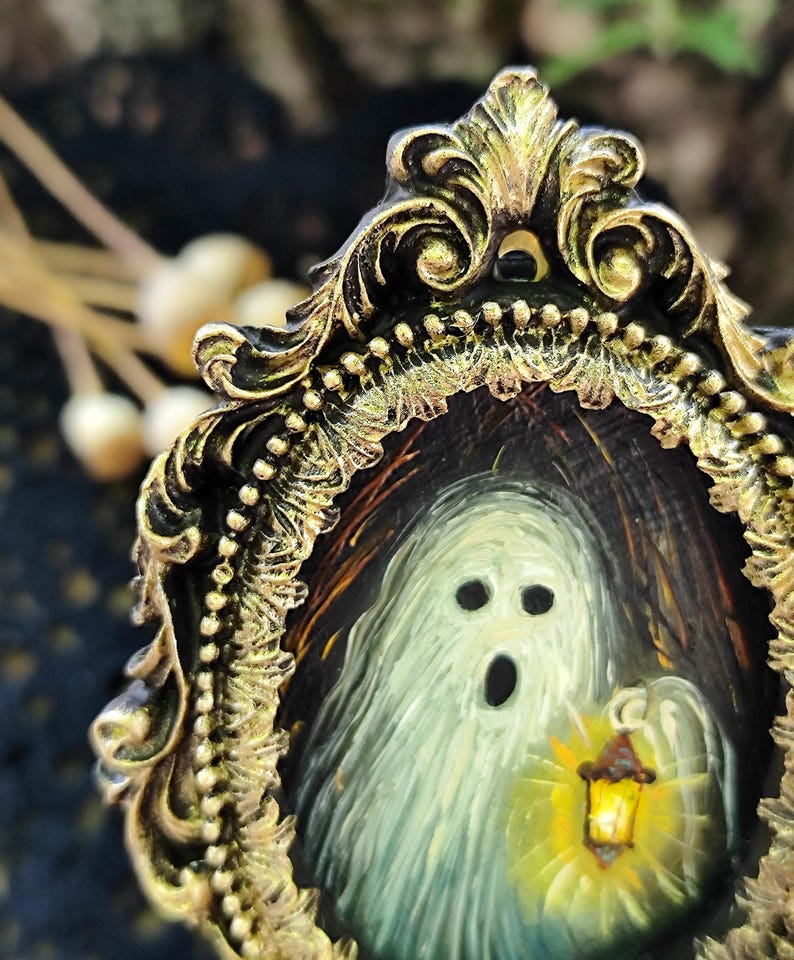 May include: A small painting of a ghost with a lantern, framed in an elaborate gold-coloured frame. The ghost is rendered in white and grey, with a black open mouth and eye sockets. The frame features detailed, decorative elements.